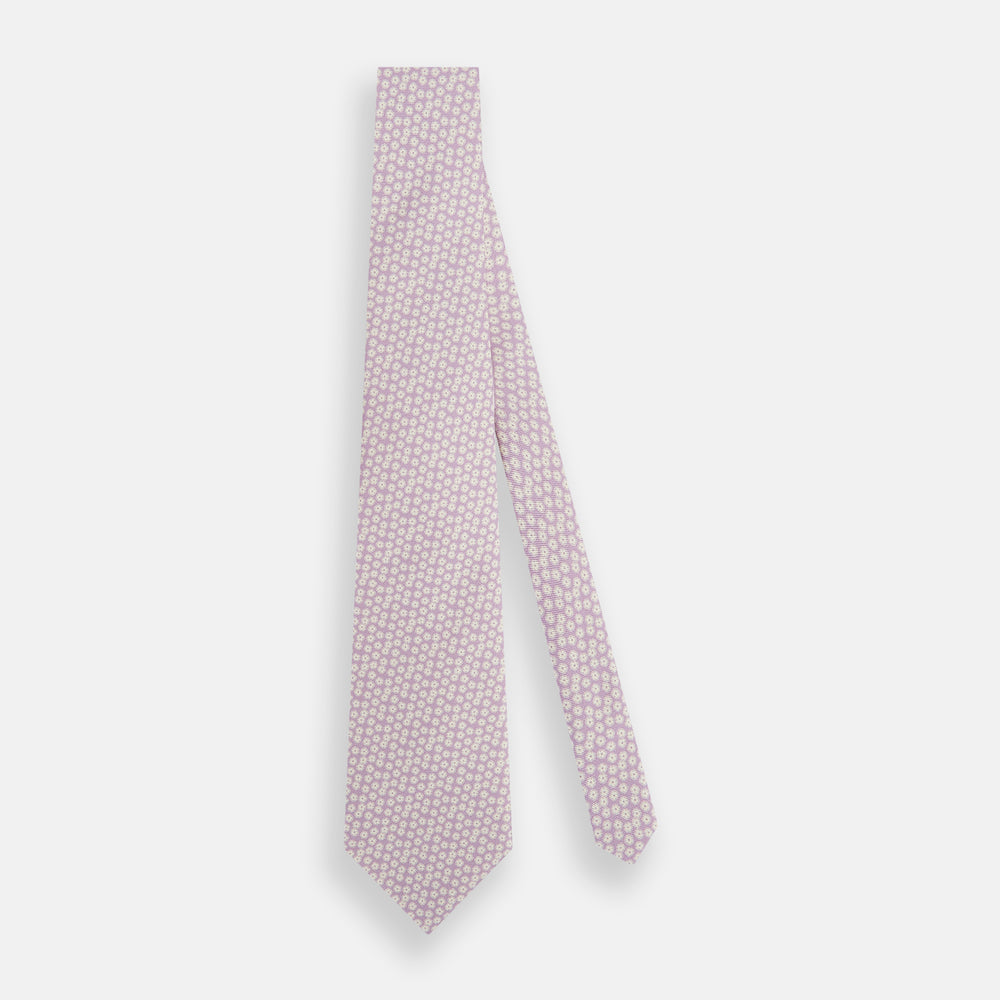 The Turnbull & Asser Lilac Flower Patterned Silk Tie features a light pink hue with a repeating white floral motif, shown rolled up on a plain white background.