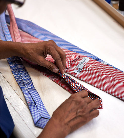 Close-up of a persons hands folding and working on patterned neckties on a white surface, with several red and blue ties laid out in the background.