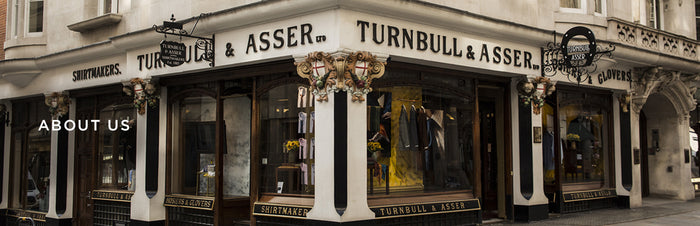 A street corner view of the Turnbull & Asser shop, a historic shirtmaker, with large windows displaying clothing and ornate architectural details above the entrance. The words ABOUT US appear on the left.