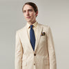 View 7 of product A man in a Turnbull & Asser Beige Cotton Barrington Blazer, light yellow shirt, blue patterned tie, and dark trousers stands against a plain light background, with a pocket square visible and a neutral expression.