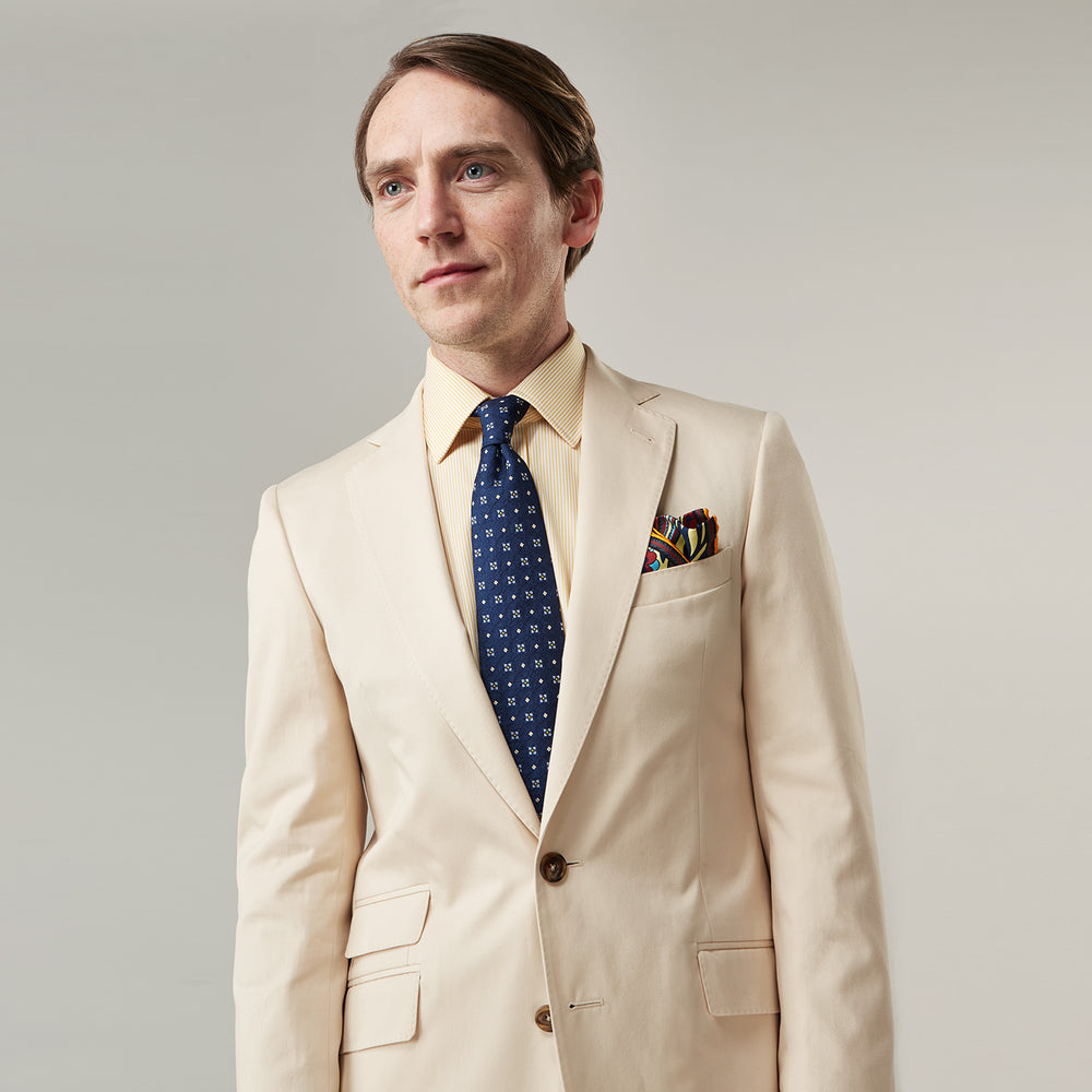 A man in a Turnbull & Asser Beige Cotton Barrington Blazer, light yellow shirt, blue patterned tie, and dark trousers stands against a plain light background, with a pocket square visible and a neutral expression.
