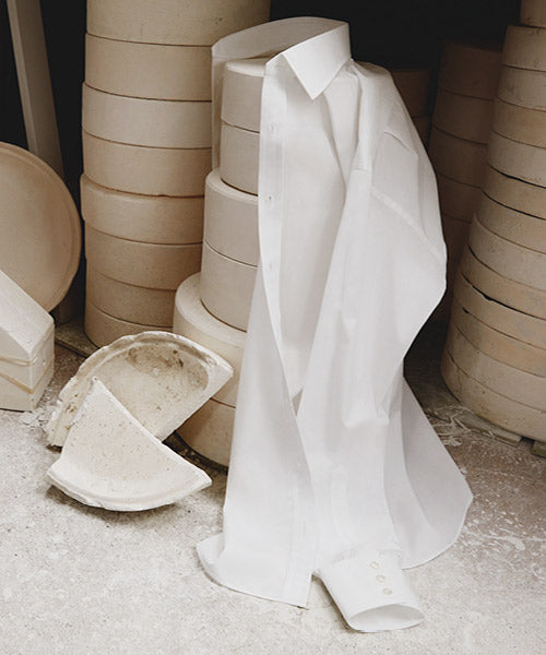 A crisp white dress shirt draped over a stack of round, beige ceramic molds, with broken ceramic pieces on the floor nearby. The scene is set against a neutral, industrial background.