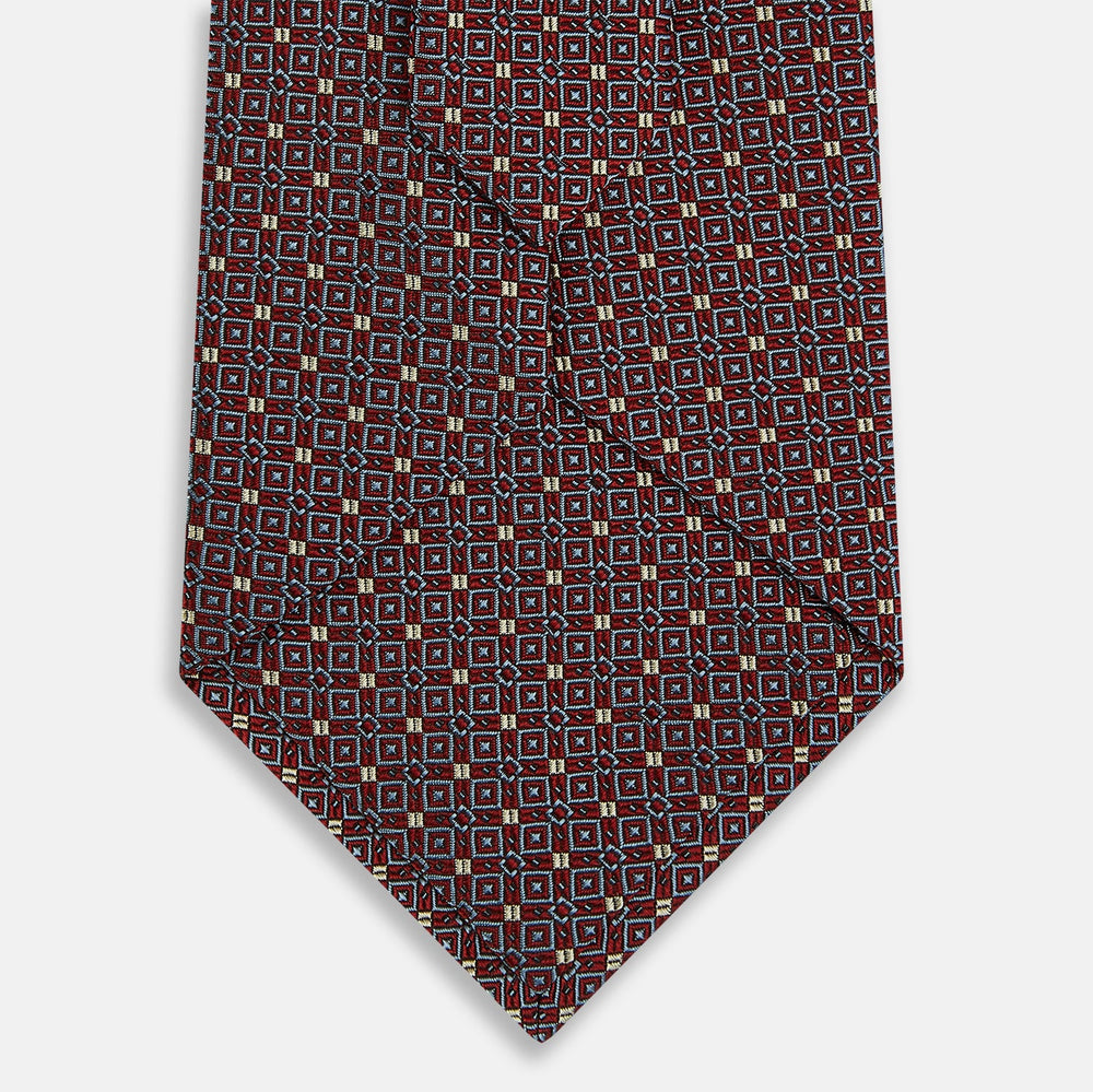 The Turnbull & Asser Burgundy Geometric Silk Tie, crafted in Britain, features a repeating pattern of light blue and white squares and diamonds. The neatly rolled tie is shown against a plain white background.