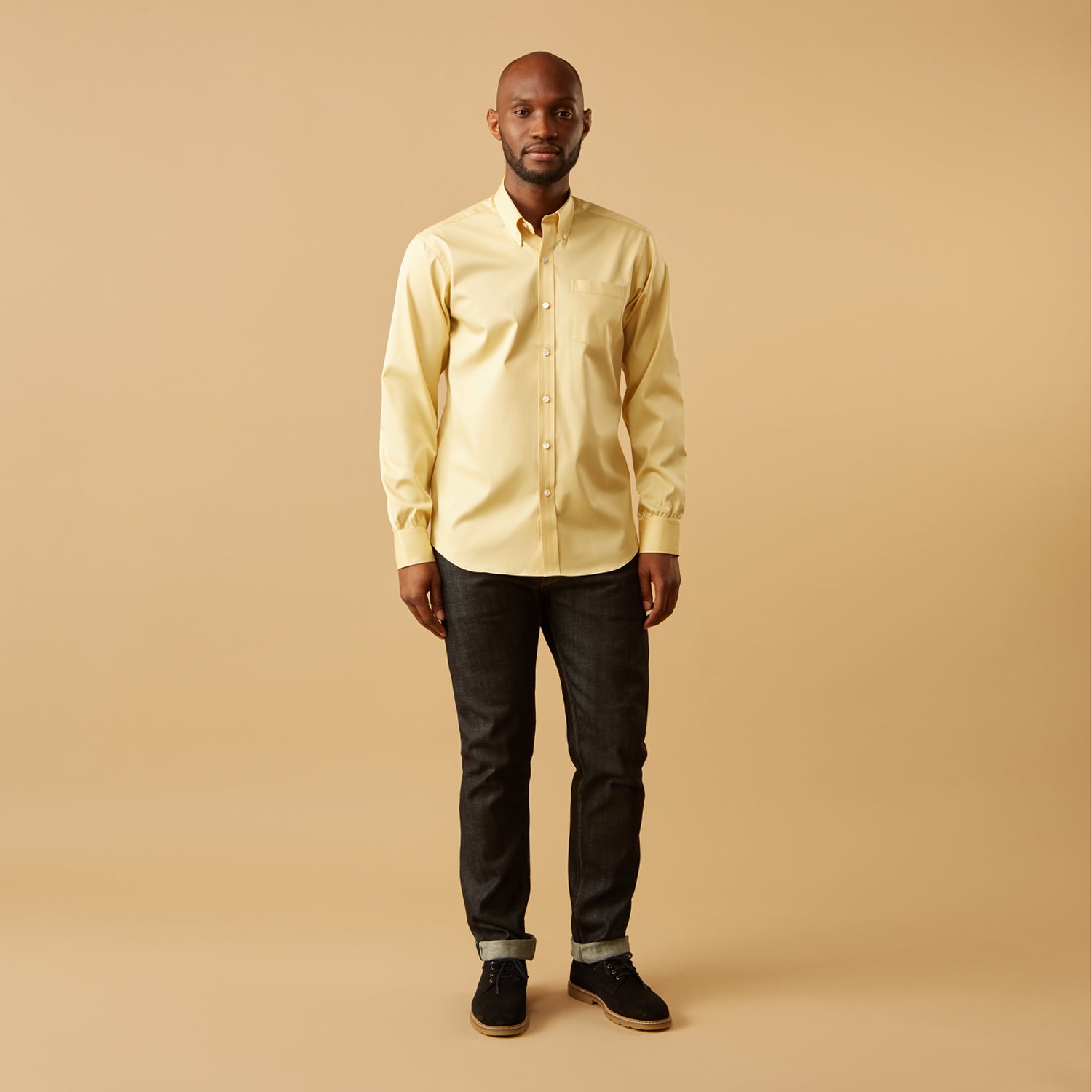 View 1 of product A man stands facing forward against a beige background, wearing the Turnbull & Asser Yellow Piece Dyed Weekend Fit Suffolk Shirt with a Dorset collar, paired with dark jeans and black shoes.