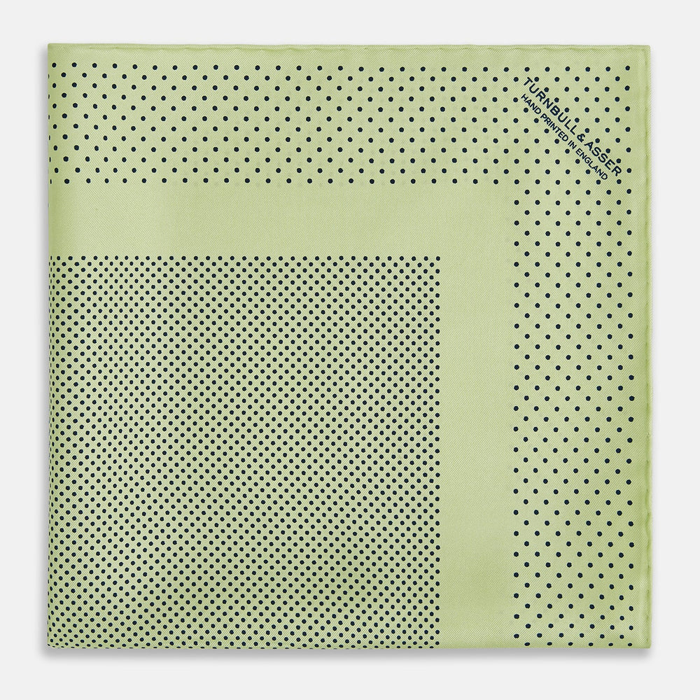 The Melon Green Dot Silk Pocket Square by Turnbull & Asser is handmade in England from silk, featuring small black polka dots. Shown folded with a turned-up corner, it displays both sides of the light green fabric.