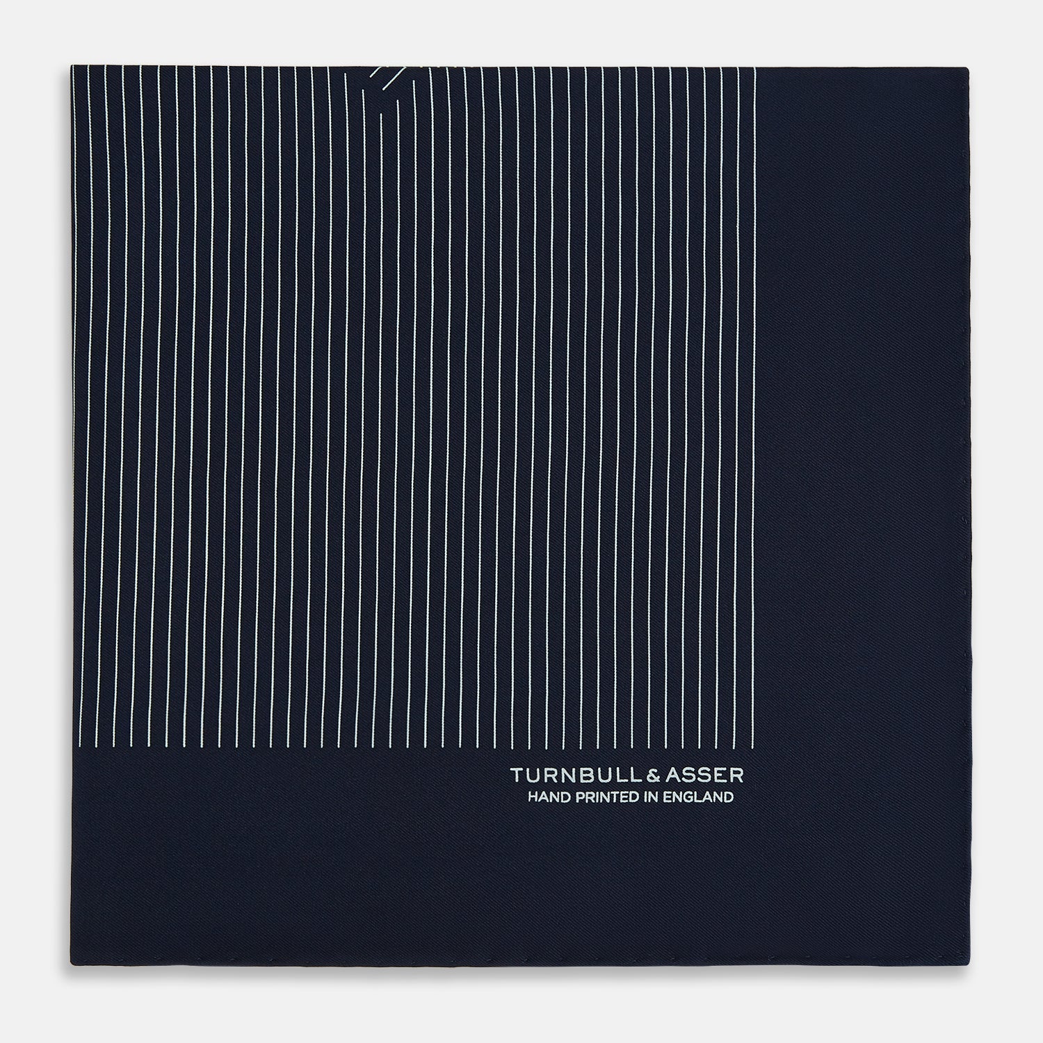 View 4 of product The Navy and White Stripe Silk Pocket Square by Turnbull & Asser features thin white vertical stripes and "TURNBULL & ASSER HAND PRINTED IN ENGLAND" in one corner, with a folded edge showing the reverse of this elegant silk accessory.