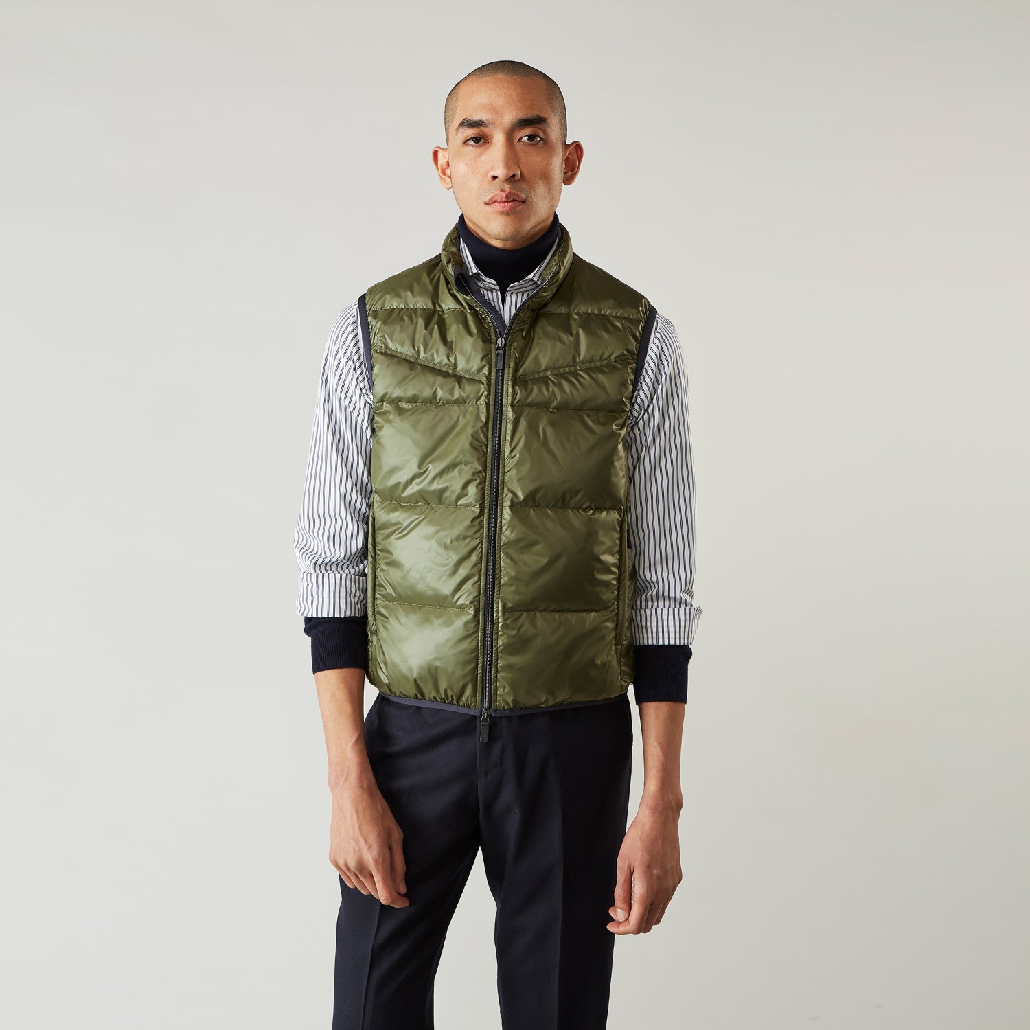 View 3 of product A man stands against a plain background wearing the Turnbull & Asser Green Peyton Sealup Quilted Gilet over a striped shirt, dark trousers, and white sneakers, with his hands in his pockets and looking slightly to the side.