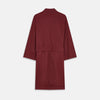 View 5 of product A person stands indoors by a window, wearing the Turnbull & Asser Burgundy Cotton-Cashmere Kimono Gown over striped pajamas, with matching slippers, next to cream curtains and a wooden chair against a pale wall.