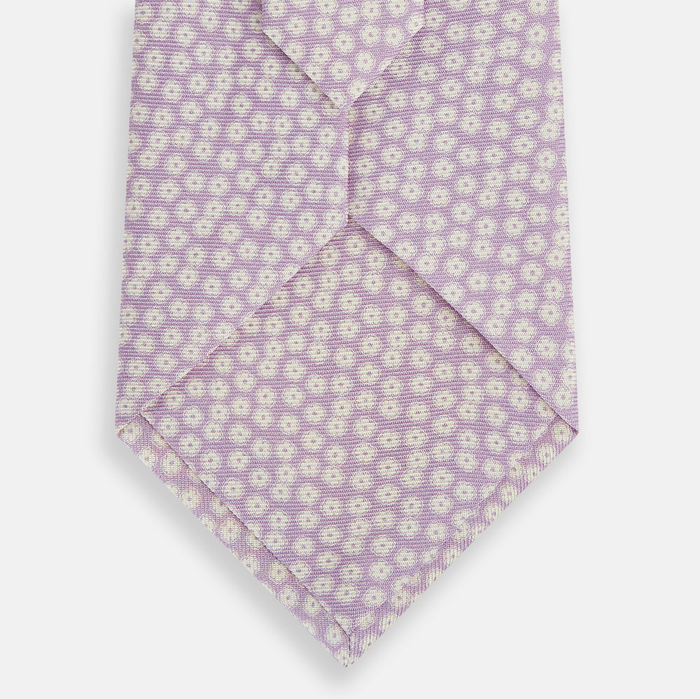 The Turnbull & Asser Lilac Flower Patterned Silk Tie features a light pink hue with a repeating white floral motif, shown rolled up on a plain white background.