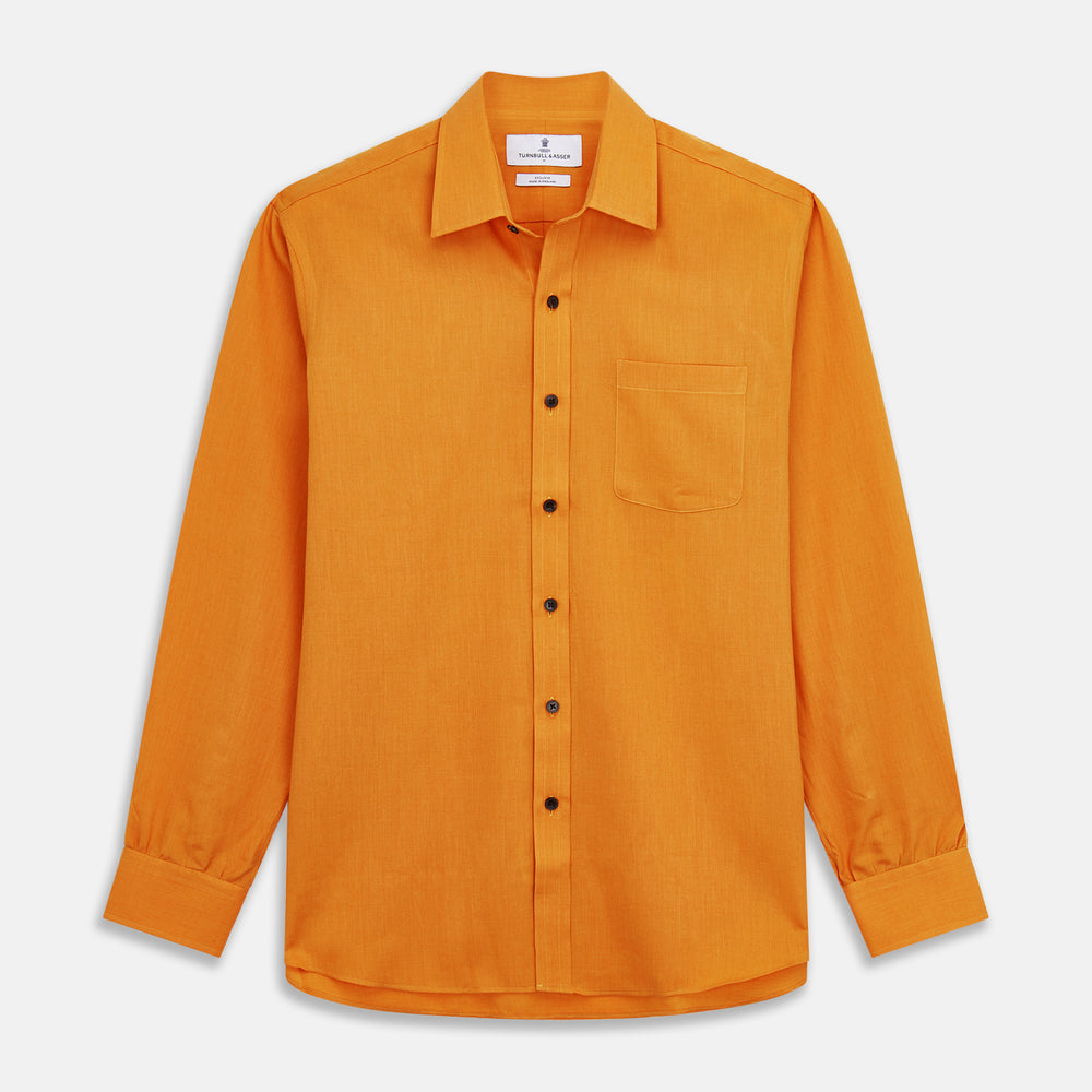 A man stands against a tan background, wearing a Turnbull & Asser Orange Weekend Fit Cotton & Wool Shirt with Derby Collar and 1 Button Cuffs, tucked into dark blue dress pants with a black belt and shoes. He has one hand in his pocket.