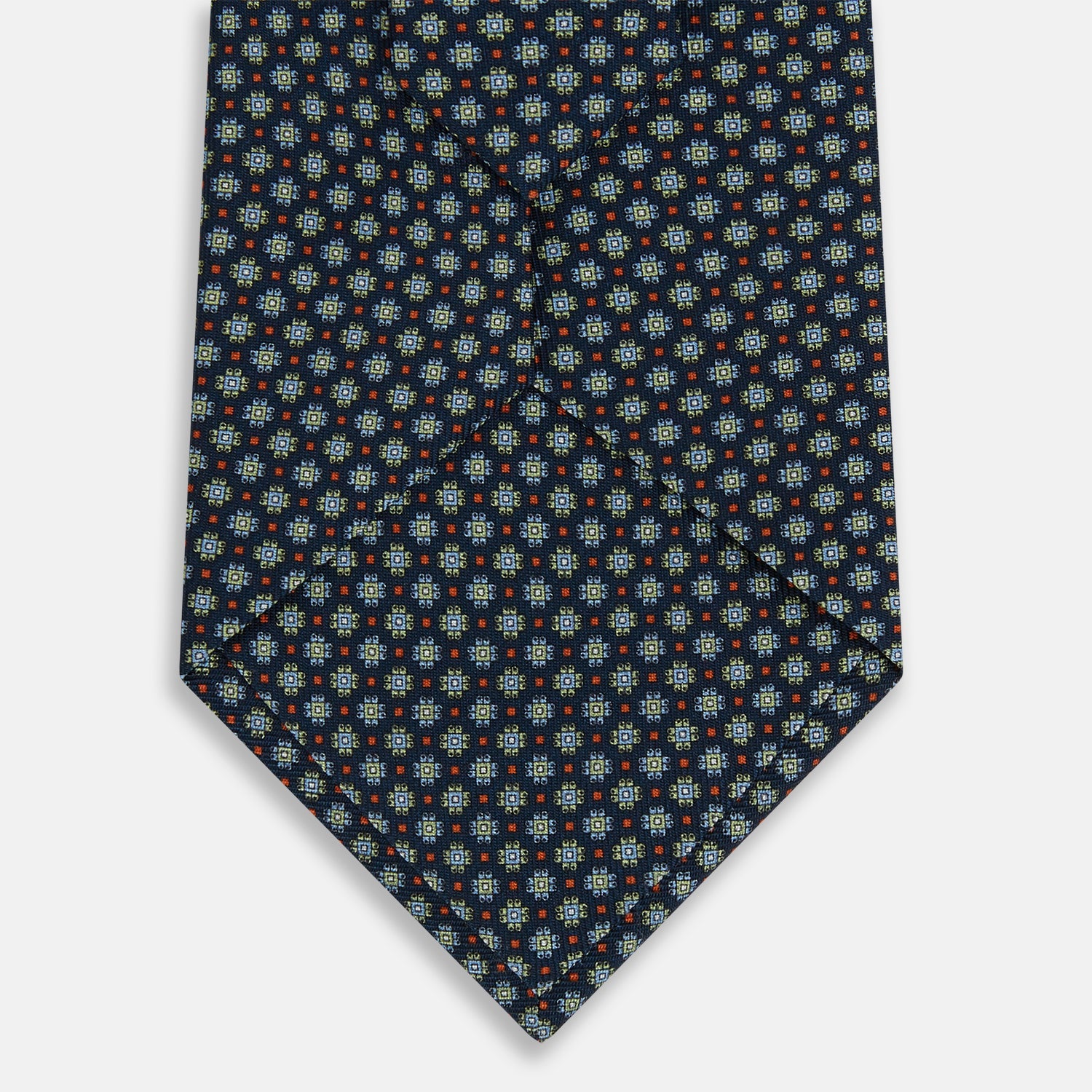 View 3 of product The Turnbull & Asser Navy Geometric Floral Silk Tie features a dark blue background with a repeating small geometric pattern in light blue, yellow, and red, shown rolled up on a plain white surface.