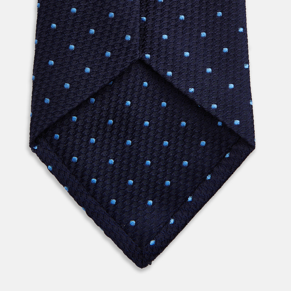 The LACE ON TONE SPOT NAVY FORD TIE by Turnbull & Asser is a rolled-up navy tie with textured fabric and an evenly spaced pattern of small light blue polka dots.