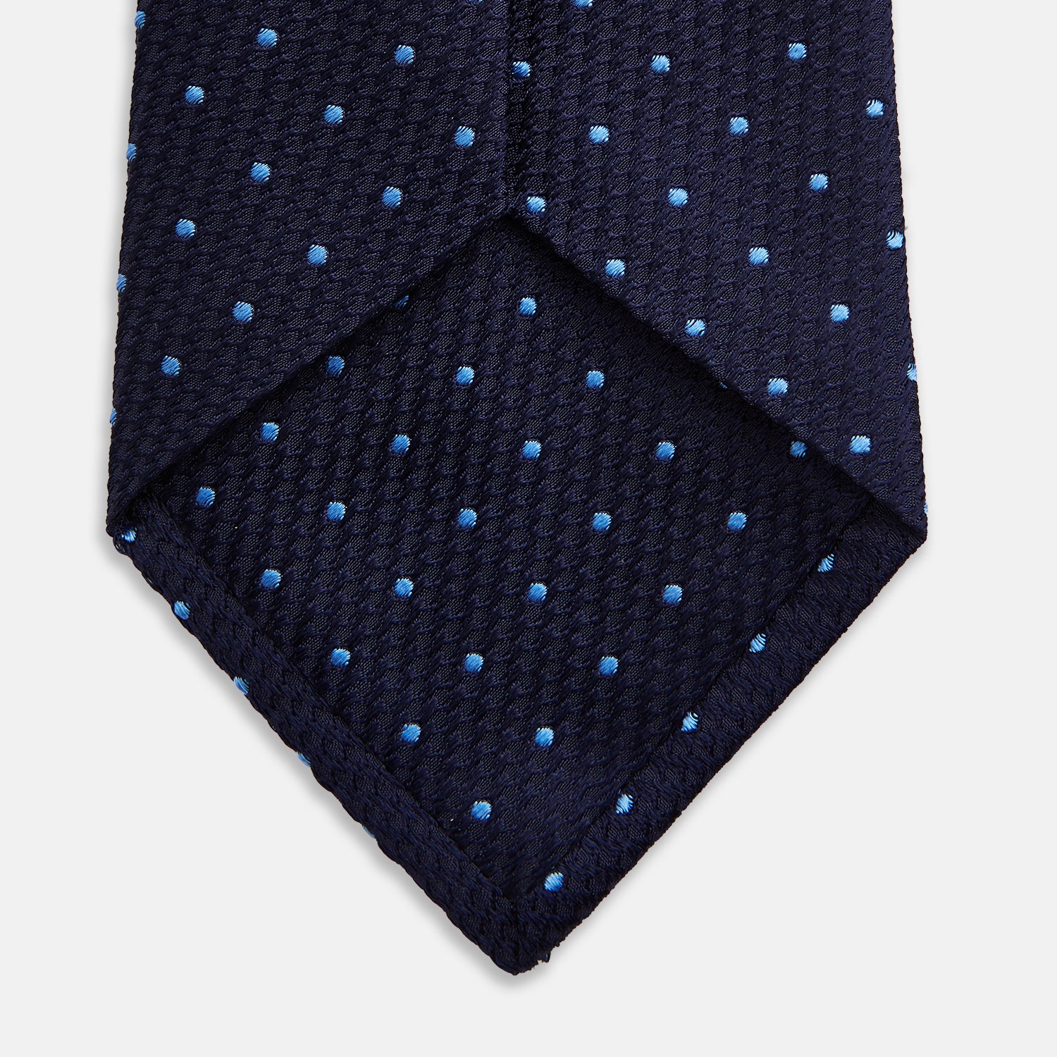 View 4 of product The LACE ON TONE SPOT NAVY FORD TIE by Turnbull & Asser is a rolled-up navy tie with textured fabric and an evenly spaced pattern of small light blue polka dots.