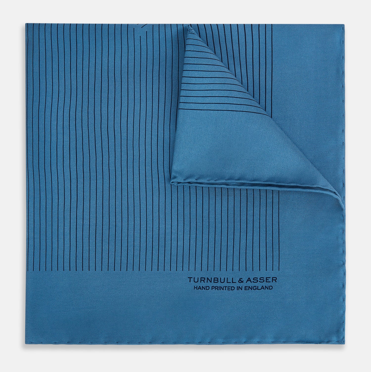View 1 of product The Blue Stripe Silk Pocket Square by Turnbull & Asser features thin vertical lines, British heritage details, and "TURNBULL & ASSER HAND PRINTED IN ENGLAND" printed on the fabric, with one corner folded to reveal the reverse side.