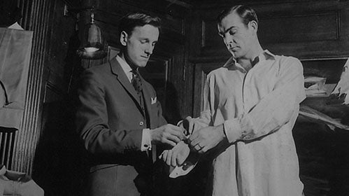 Two men in a dimly lit room; one is wearing a suit and helping the other, who is in a white shirt, put on or adjust something on his wrist. The atmosphere appears serious and formal.