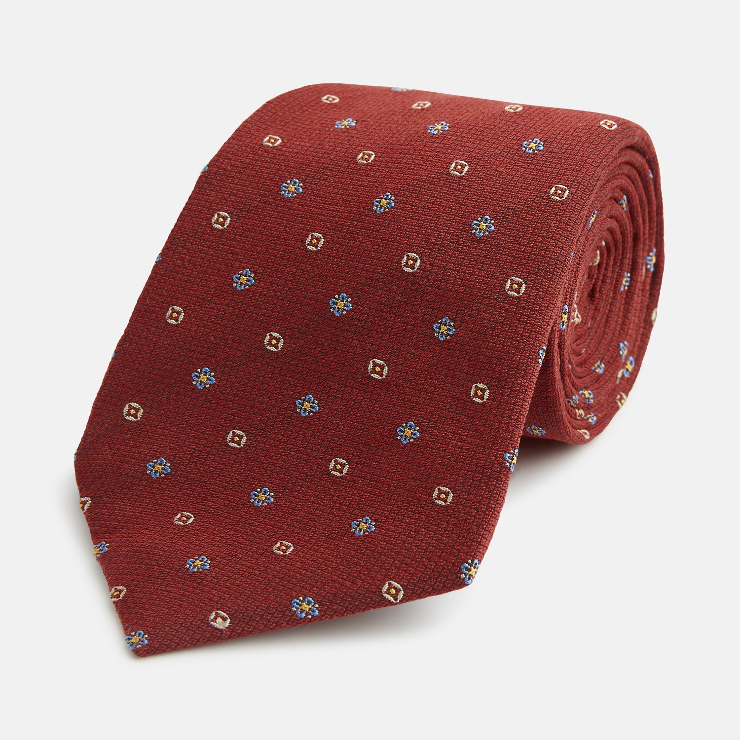View 1 of product A Turnbull & Asser Red Motif Silk Tie featuring small blue and yellow floral designs with brown geometric shapes is displayed rolled up on a plain white background.