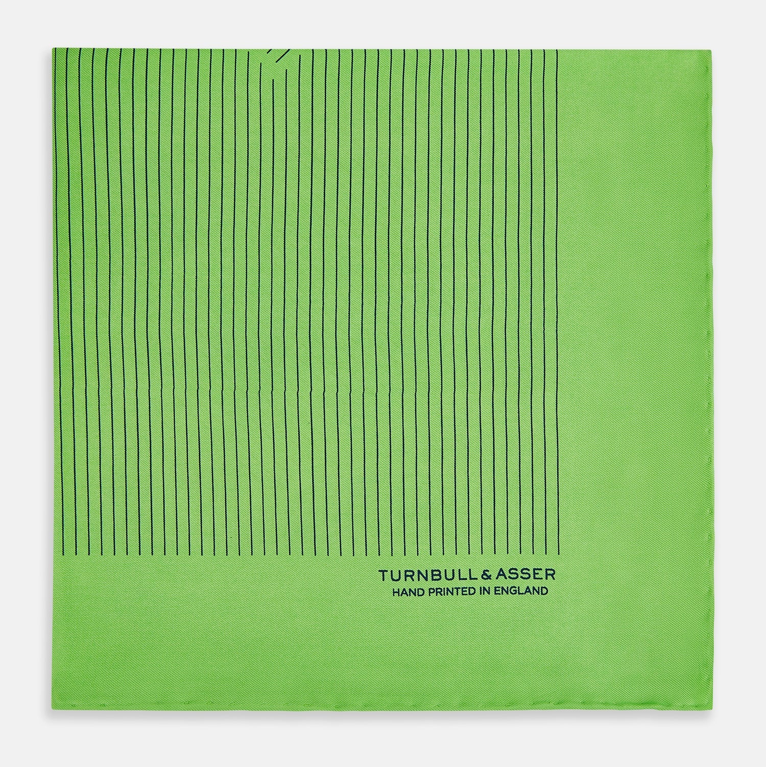 View 4 of product The Fern Green Stripe Silk Pocket Square by Turnbull & Asser features thin black vertical stripes, celebrates British heritage, and is inscribed “TURNBULL & ASSER HAND PRINTED IN ENGLAND” in one corner; one corner is folded inward.