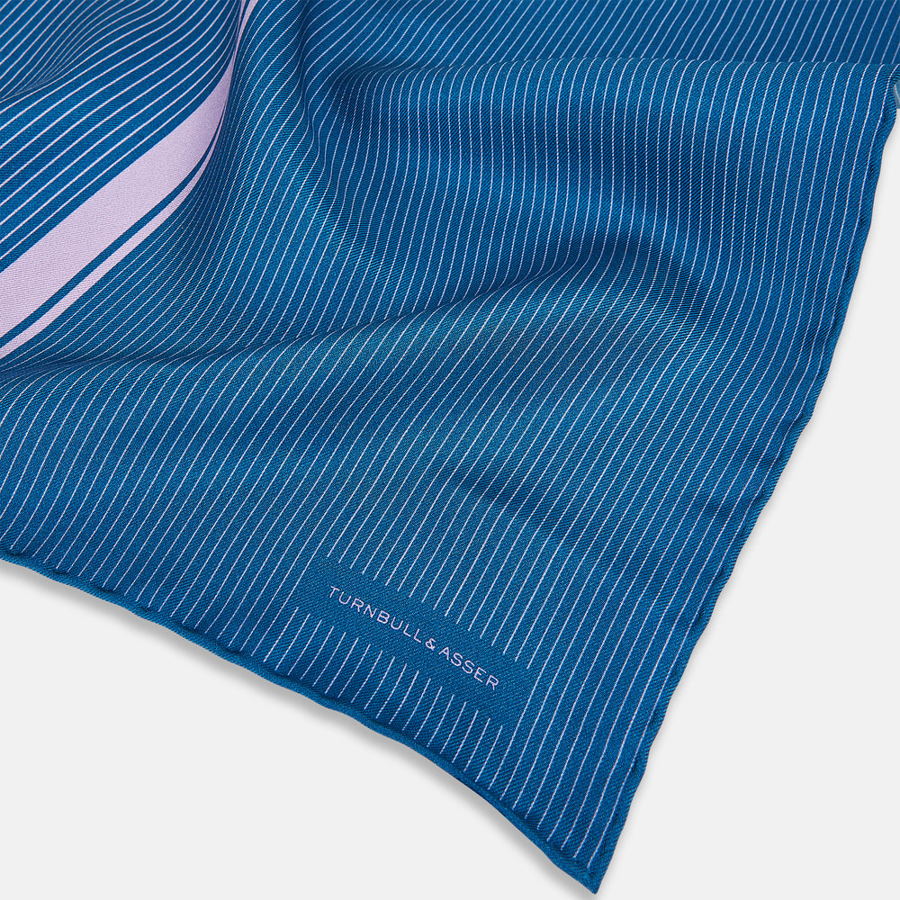 The Turnbull & Asser Blue and Lilac Stripe Silk Pocket Square features blue silk with white pinstripes, a bold lilac stripe near the edge, a turned-up corner revealing the reverse, and the brand name displayed in the bottom right.