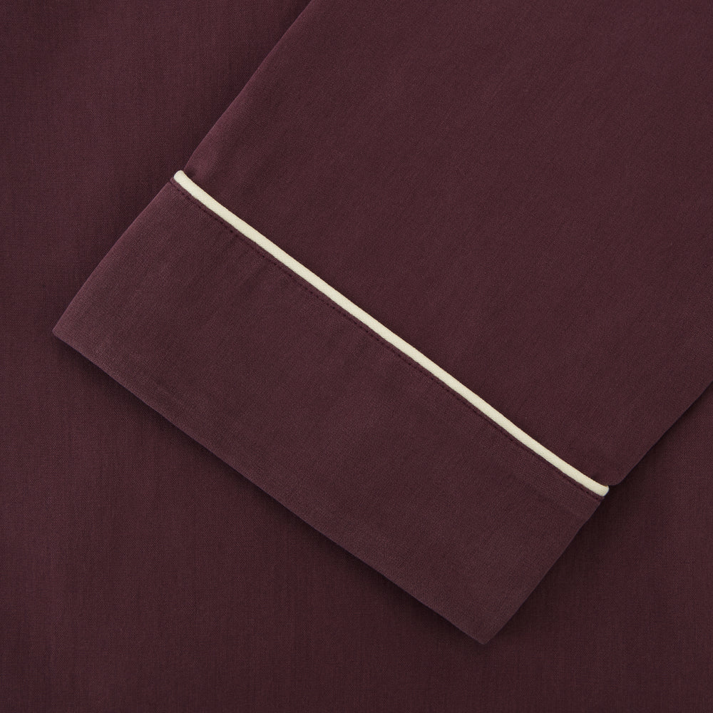 The Turnbull & Asser Plain Plum Silk Modern Pyjama Set, made in England, features white piping, a button-up long-sleeve top with three pockets, and matching pants. The pyjamas are shown against a white background.