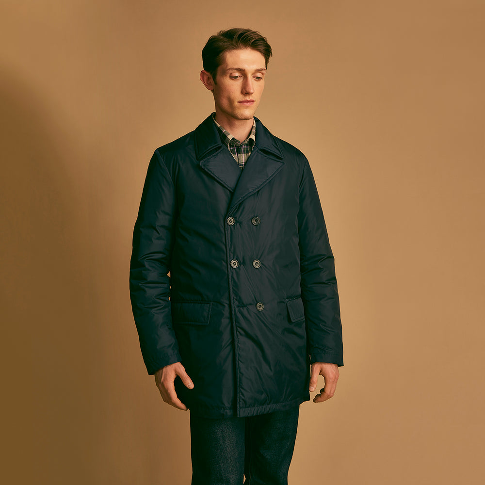 A man stands against a beige background, wearing the Turnbull & Asser Navy Burrows Double Breasted Coat over a plaid shirt and mauve cardigan, paired with dark rolled-up jeans and black shoes, looking directly at the camera with a neutral expression.