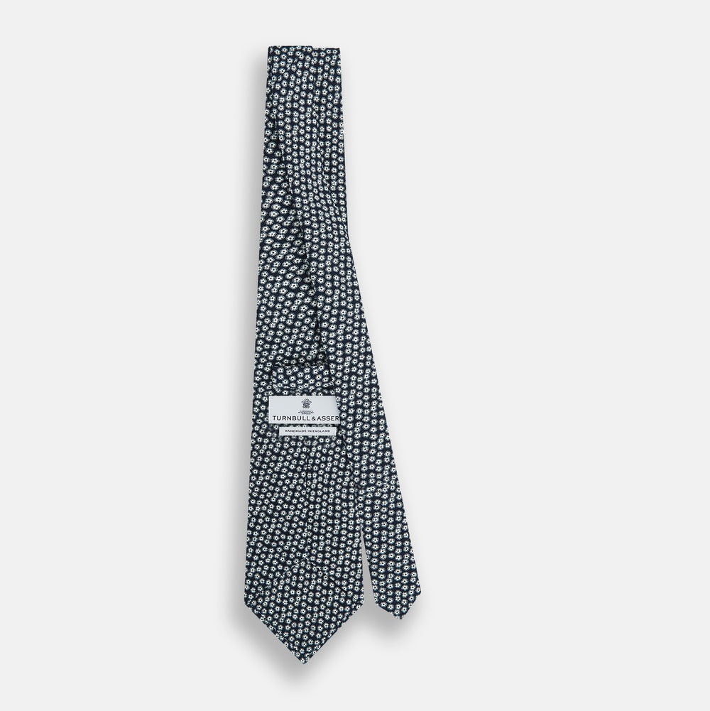 The Turnbull & Asser Navy Flower Patterned Silk Tie is shown rolled up, displaying a repeating floral design with small white and light blue circles on navy, set against a plain white background.
