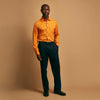 View 1 of product A man stands against a tan background, wearing a Turnbull & Asser Orange Weekend Fit Cotton & Wool Shirt with Derby Collar and 1 Button Cuffs, tucked into dark blue dress pants with a black belt and shoes. He has one hand in his pocket.