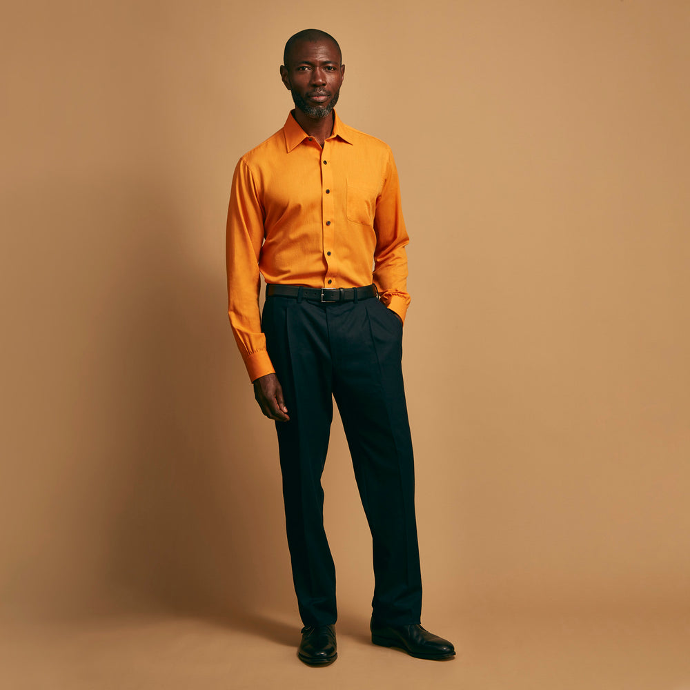 A man stands against a tan background, wearing a Turnbull & Asser Orange Weekend Fit Cotton & Wool Shirt with Derby Collar and 1 Button Cuffs, tucked into dark blue dress pants with a black belt and shoes. He has one hand in his pocket.