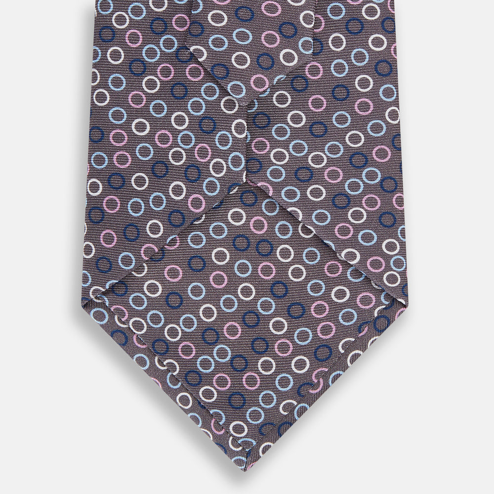 The Turnbull & Asser Purple and Blue Circle Ash Silk Tie features small blue, pink, and white circles on a dark purple background, adding an elegant touch when displayed on a light surface.