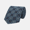 View 1 of product The Jacquard Silk Geometric Tie by Turnbull & Asser features a navy blue base with light blue and white star and circle motifs, shown rolled on a white background.