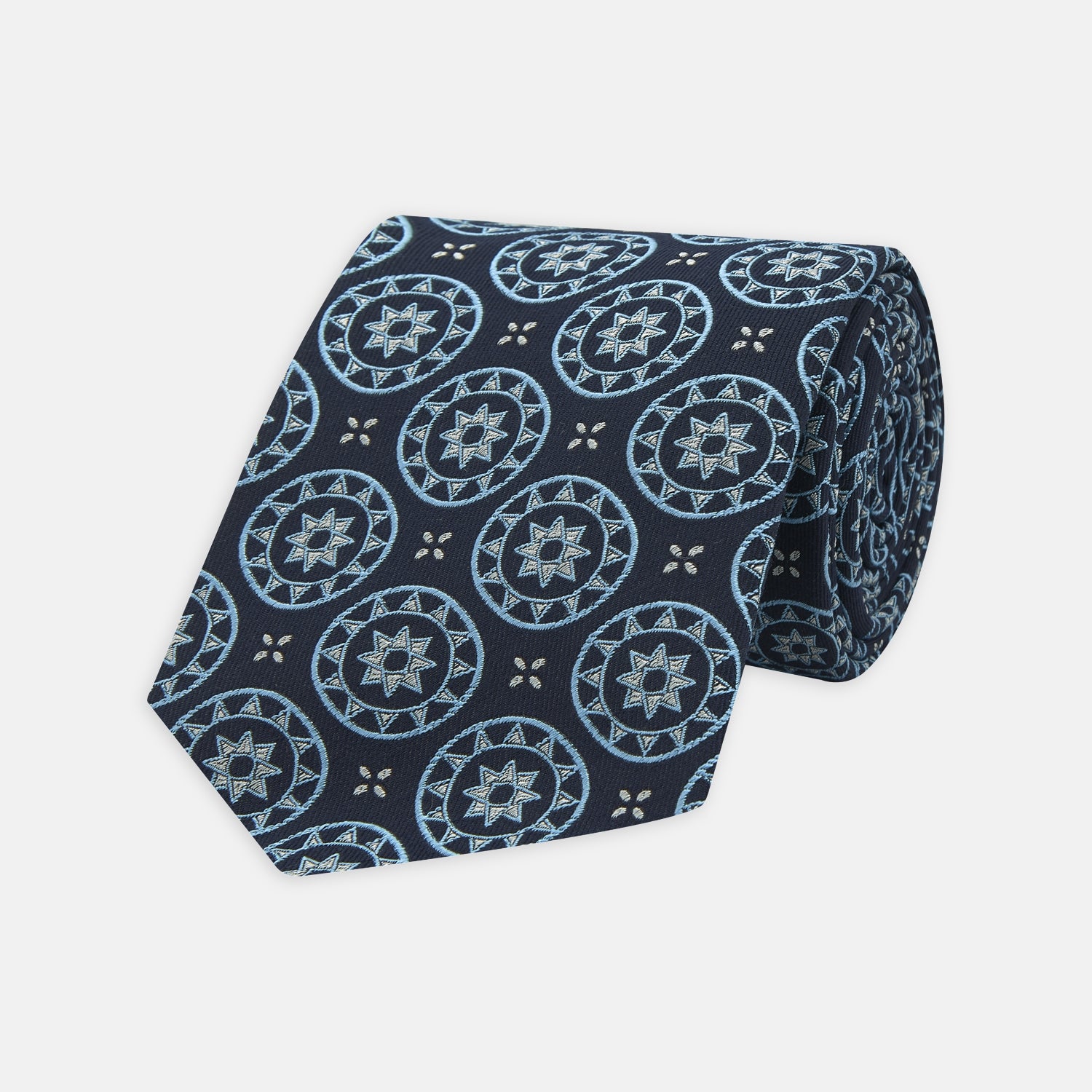 View 1 of product The Jacquard Silk Geometric Tie by Turnbull & Asser features a navy blue base with light blue and white star and circle motifs, shown rolled on a white background.