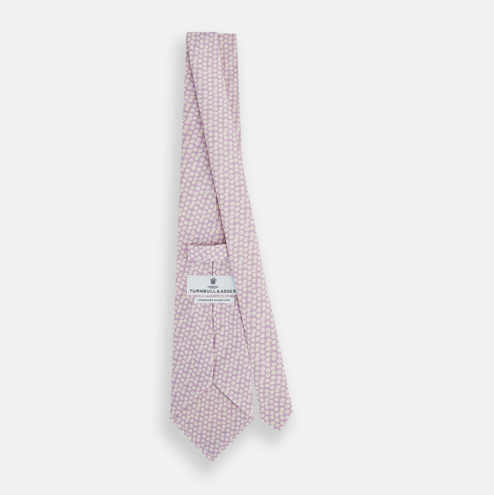 The Turnbull & Asser Lilac Flower Patterned Silk Tie features a light pink hue with a repeating white floral motif, shown rolled up on a plain white background.