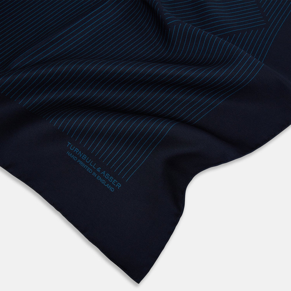 The Navy and Blue Stripe Silk Pocket Square by Turnbull & Asser features vertical blue pinstripes, is crafted from silk with hand-rolled edges, and is finished with the brand's logo printed in blue at the lower right corner.