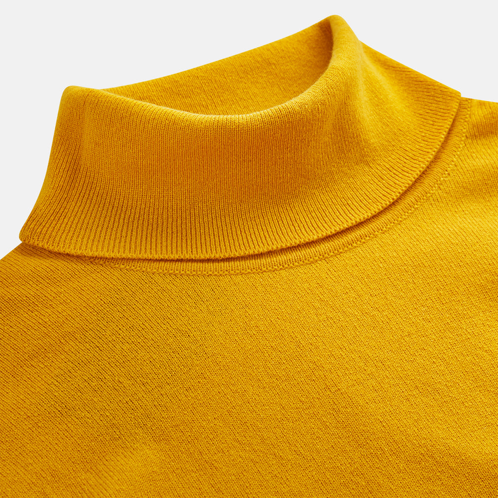 A man with short hair and a beard wears the Gold Aubrey Cashmere Roll Neck Jumper by Turnbull & Asser with dark pants, standing against a plain beige background and calmly looking at the camera, highlighting this stylish autumn sweater look.