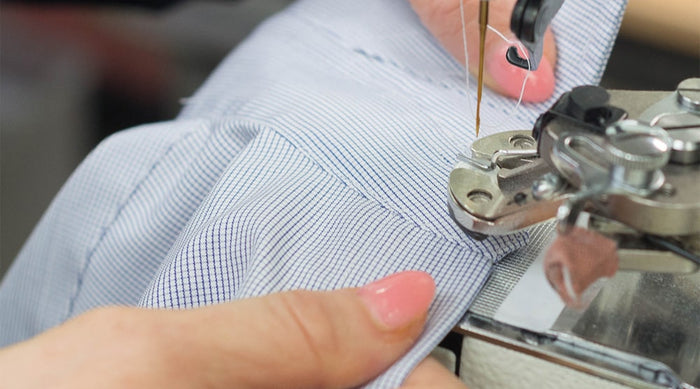 Close-up of hands guiding light blue striped fabric through a sewing machine, showing the needle stitching the material. The person has pink nail polish.