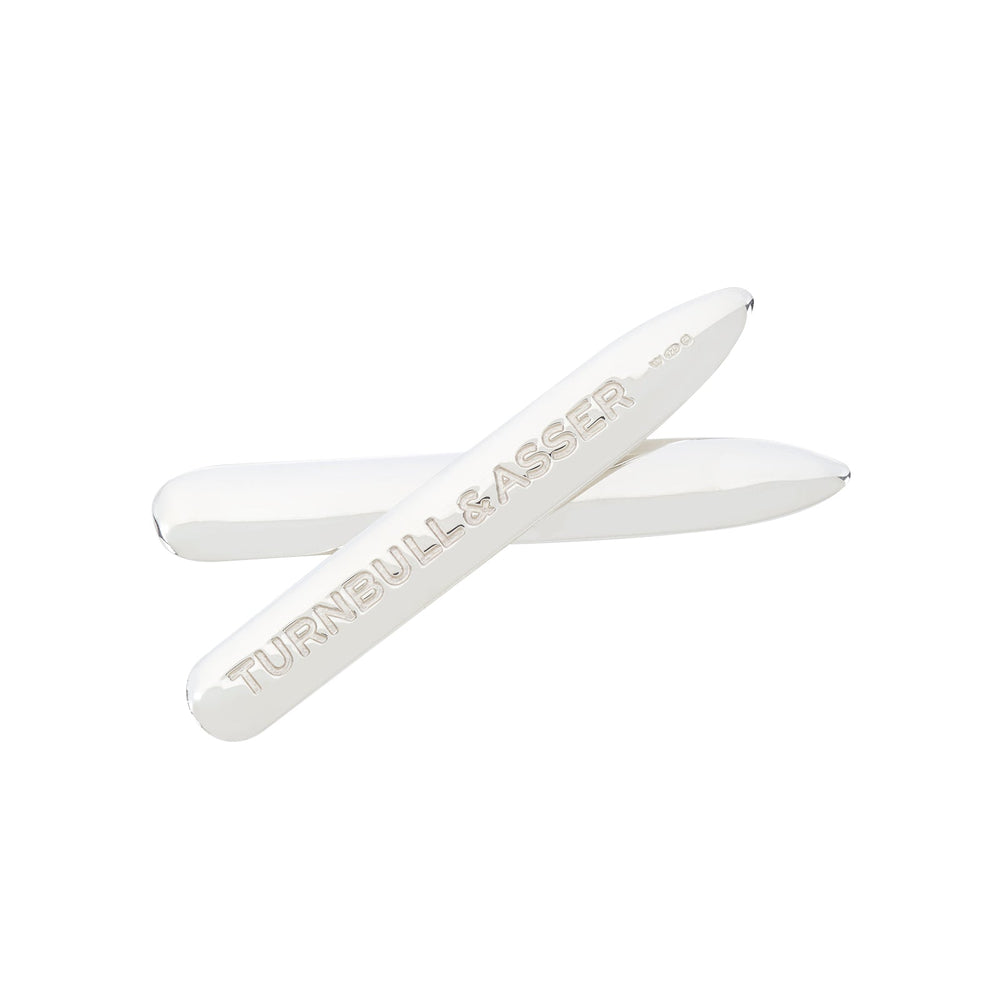 Two Short Sterling Silver Collar Stays by Turnbull & Asser are displayed on a white background; one is engraved with the brand name, while the other is plain. Both have rounded tips and a polished, reflective finish.