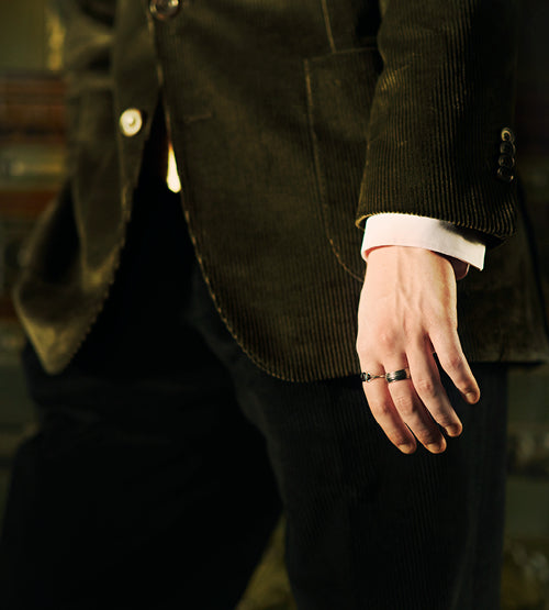 A person wearing a dark corduroy blazer and pants stands with their hand visible, showing a ring on one finger. The background is blurred.