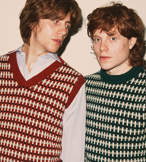 Two people with reddish-brown hair wear patterned knit vests over collared shirts. One vest is red and white, the other green and white. Both stand closely against a neutral background, with one person looking at the other.