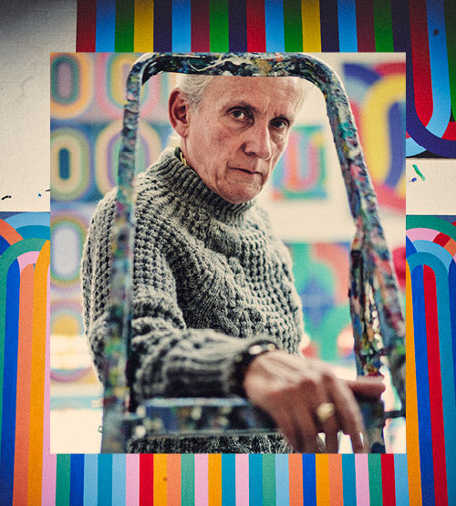 A person with short gray hair, wearing a chunky knit sweater, looks seriously at the camera through a paint-splattered metal frame, with colorful geometric patterns in the background.