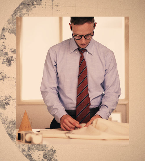 A man wearing glasses, a light blue dress shirt, and a red striped tie concentrates while working with fabric and tools on a table in a softly lit room.