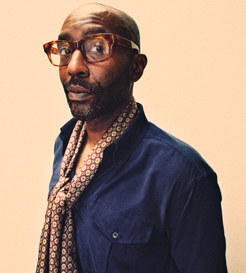 A man with a bald head and beard wearing large brown glasses, a navy blue shirt, and a patterned scarf, stands against a plain light background, looking confidently at the camera.
