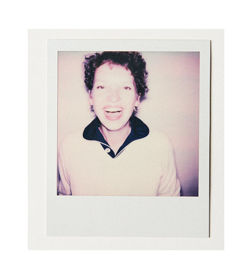 A person with short curly hair smiles widely at the camera, wearing a light-colored sweatshirt with a dark collar, against a plain light background in a Polaroid-style photo.