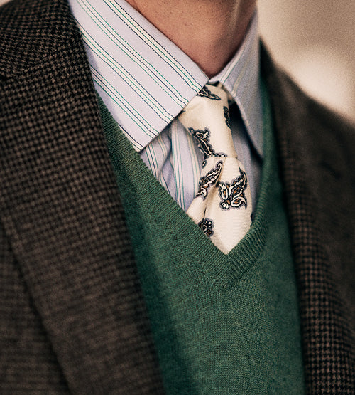 Close-up of a person wearing a brown checked blazer, green V-neck sweater, blue and white striped dress shirt, and a white tie with a black paisley pattern.