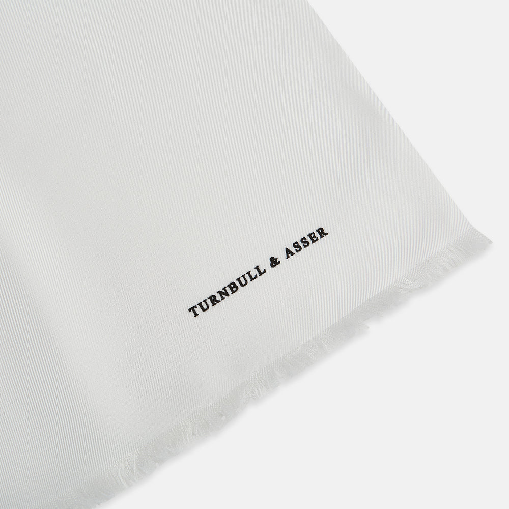 The Turnbull & Asser White Silk Scarf features fringed edges and discreet black branding in the corner, making it a refined accessory for tuxedo jackets or formal wear. Presented neatly folded on a plain white background.