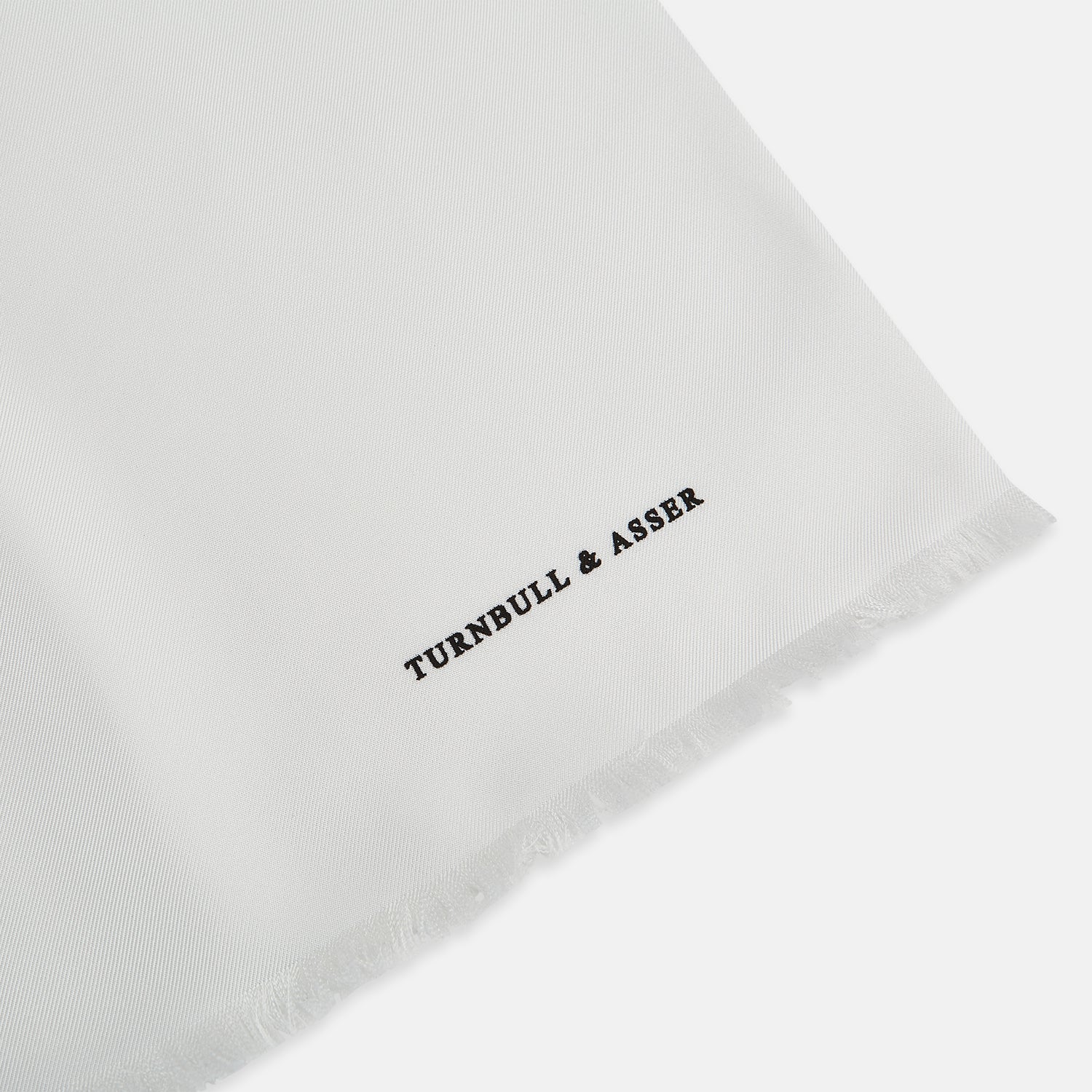 View 2 of product The Turnbull & Asser White Silk Scarf features fringed edges and discreet black branding in the corner, making it a refined accessory for tuxedo jackets or formal wear. Presented neatly folded on a plain white background.