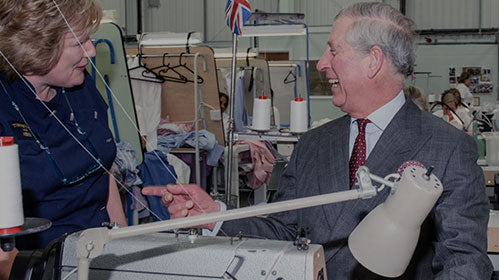 A man in a suit and a woman in a work uniform smile and talk beside a sewing machine in a busy workshop, with fabric and thread spools visible in the background.