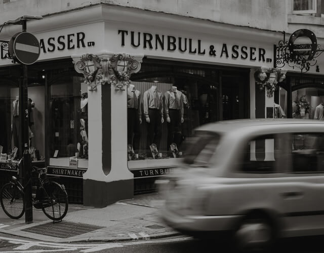 A black and white photo of a street corner with a Turnbull & Asser store, displaying shirts in the window. A moving car and a bicycle are visible in the foreground.
