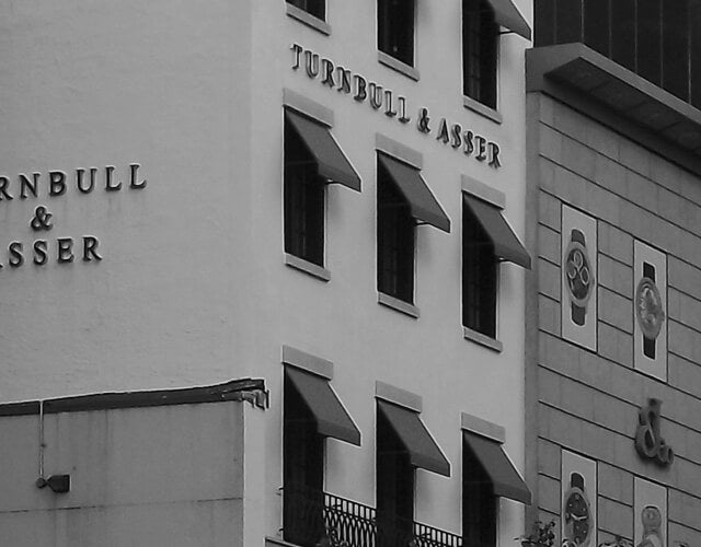 Black and white photo of a building with multiple windows and awnings, displaying the sign Turnbull & Asser on its facade. Another building with decorative clock faces is partially visible to the right.