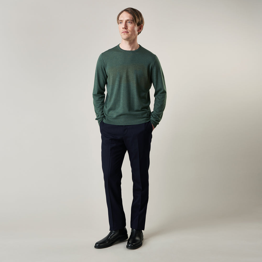 A man stands facing the camera against a plain light background, wearing the Turnbull & Asser Bottle Green Merino Kingston Crew Neck Jumper with dark pants and black shoes. His arms rest at his sides and he has a neutral expression.