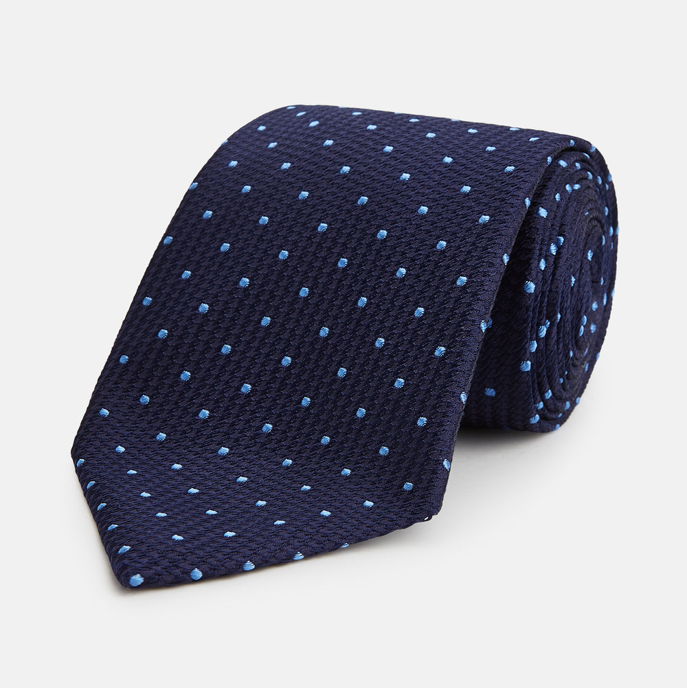 The LACE ON TONE SPOT NAVY FORD TIE by Turnbull & Asser is a rolled-up navy tie with textured fabric and an evenly spaced pattern of small light blue polka dots.