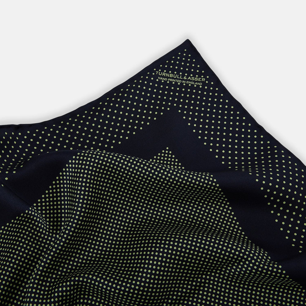 The Turnbull & Asser Navy and Melon Green Dot Silk Pocket Square features small melon green dots on navy silk with a solid border. One corner is folded over to highlight its exquisite handmade craftsmanship.